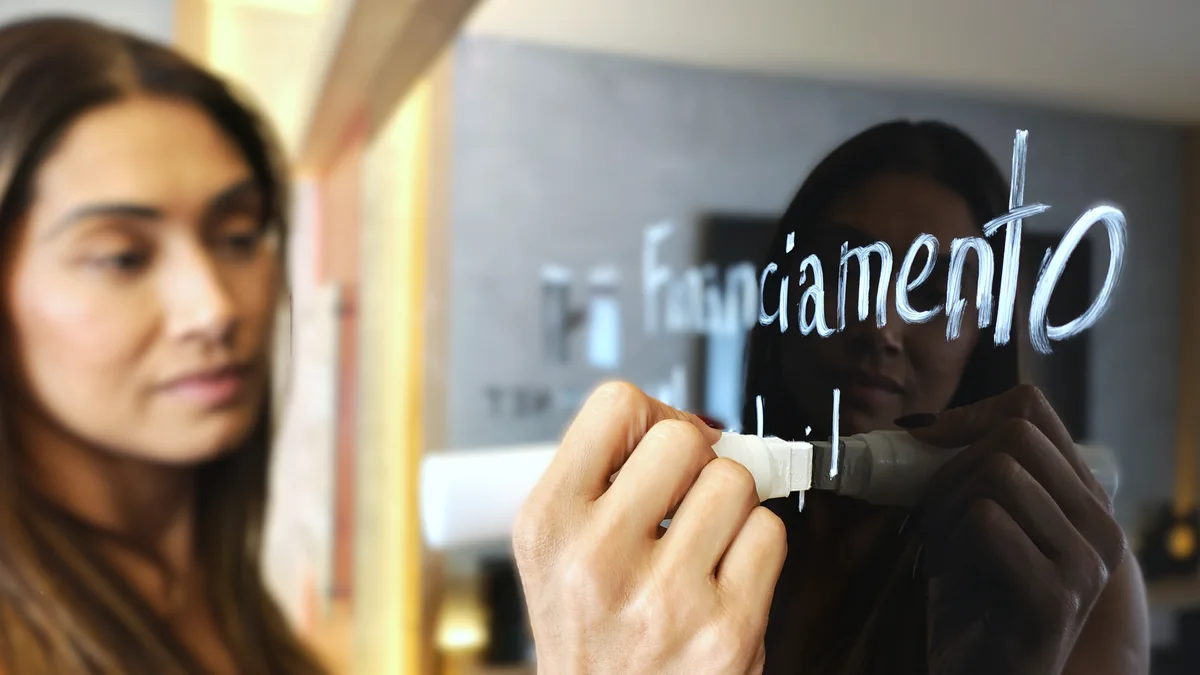 Mulher escrevendo “financiamento” em vidro, com reflexo ao fundo